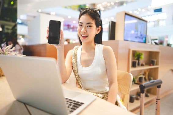 Garota da Geração Z trabalhando, segurando um telefone, com um notebook à sua frente e a alça de uma mala ao seu lado.