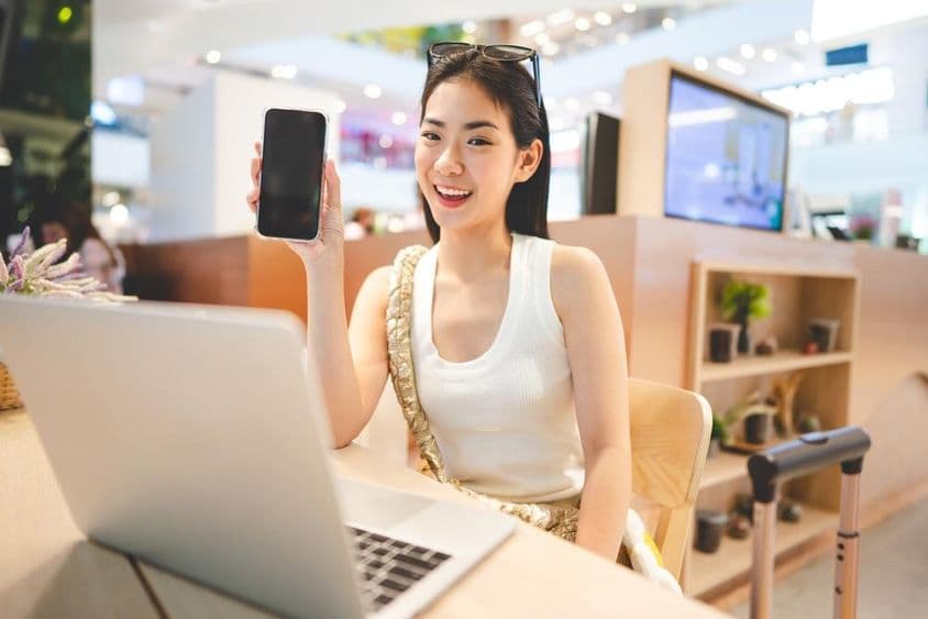 Garota da Geração Z trabalhando, segurando um telefone, com um notebook à sua frente e a alça de uma mala ao seu lado.