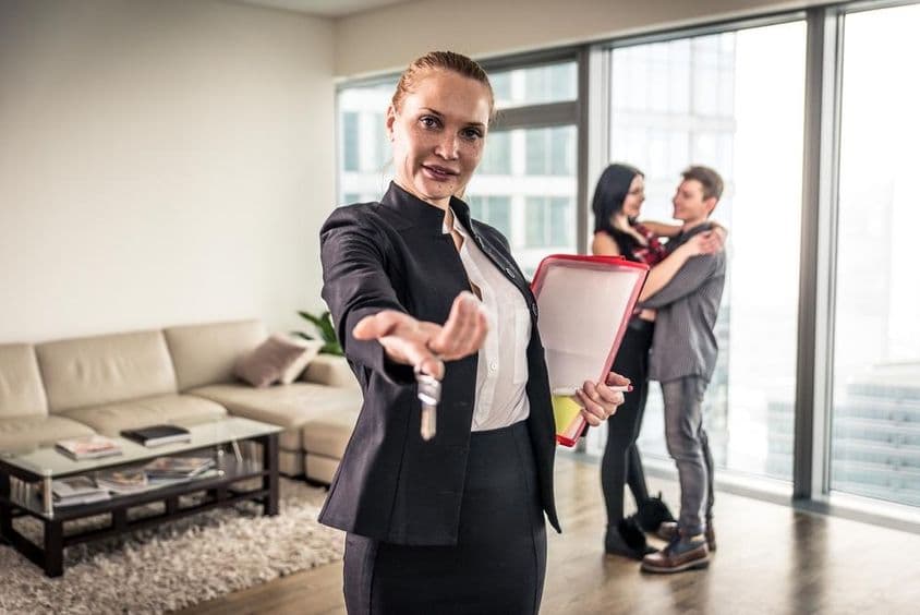 Uma corretora de imóveis acenando, um casal se abraçando ao fundo.