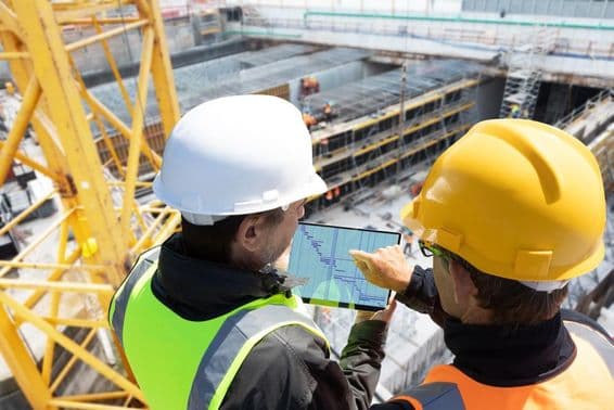 Duas pessoas com capacetes em pé em um guindaste em um canteiro de obras, conferindo planos em um tablet, com o canteiro de obras à frente delas.
