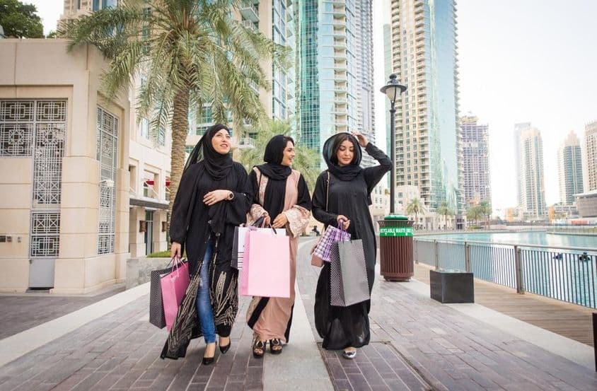 3 mulheres emiradenses caminhando com sacolas de compras após compras, com arranha-céus ao fundo.