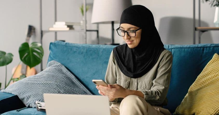 Garota árabe de hijab trabalhando em casa usando smartphone e laptop enquanto está sentada no sofá.