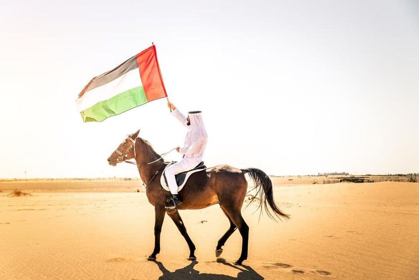 Homem do Oriente Médio em vestimenta tradicional dos Emirados montando um cavalo árabe no deserto de Dubai.