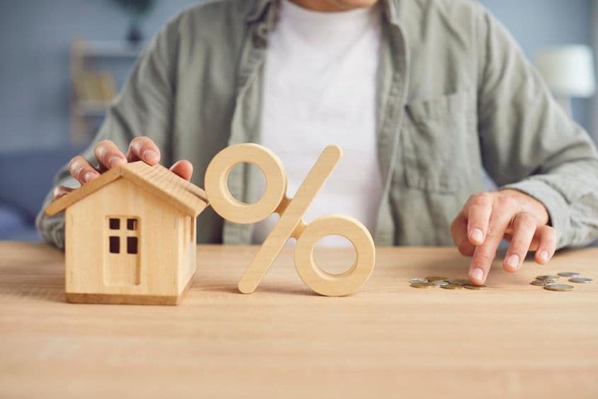 Símbolo de porcentagem em madeira e modelo de casa sobre uma mesa com um jovem contando moedas ao fundo.