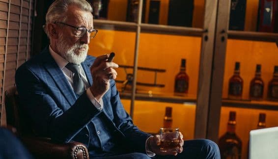 Homem idoso aposentado usando laptop, fumando, segurando licor caro.