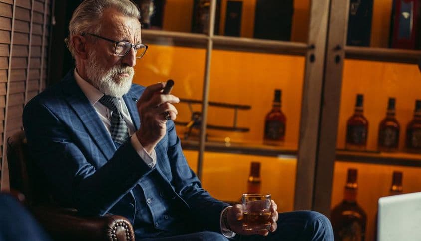 Homem idoso aposentado usando laptop, fumando, segurando licor caro.