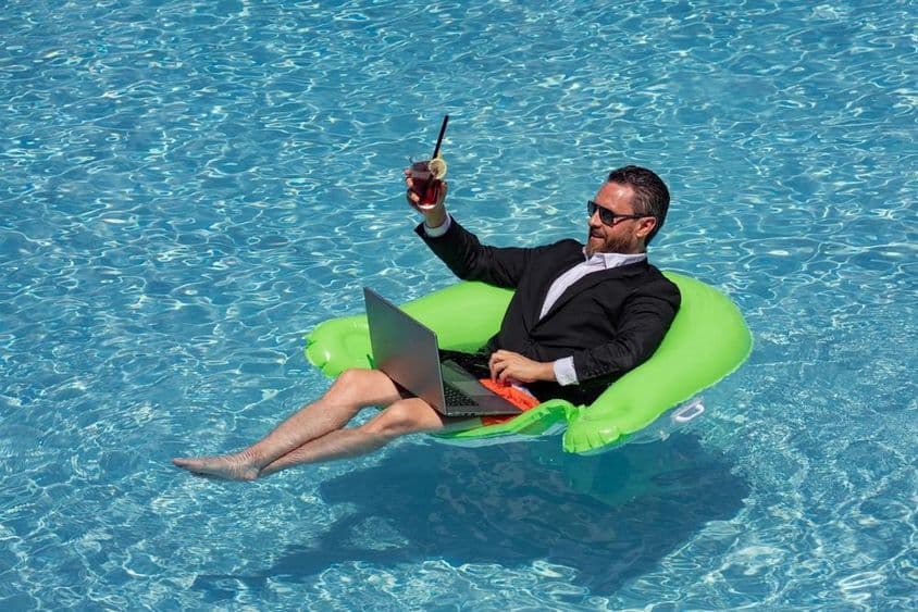 Empresário de sucesso flutuando em um terno na água da piscina, bebendo um coquetel de verão e usando um laptop na piscina.