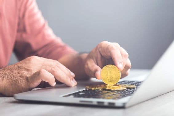 Homem segurando um Bitcoin dourado enquanto usa um laptop.