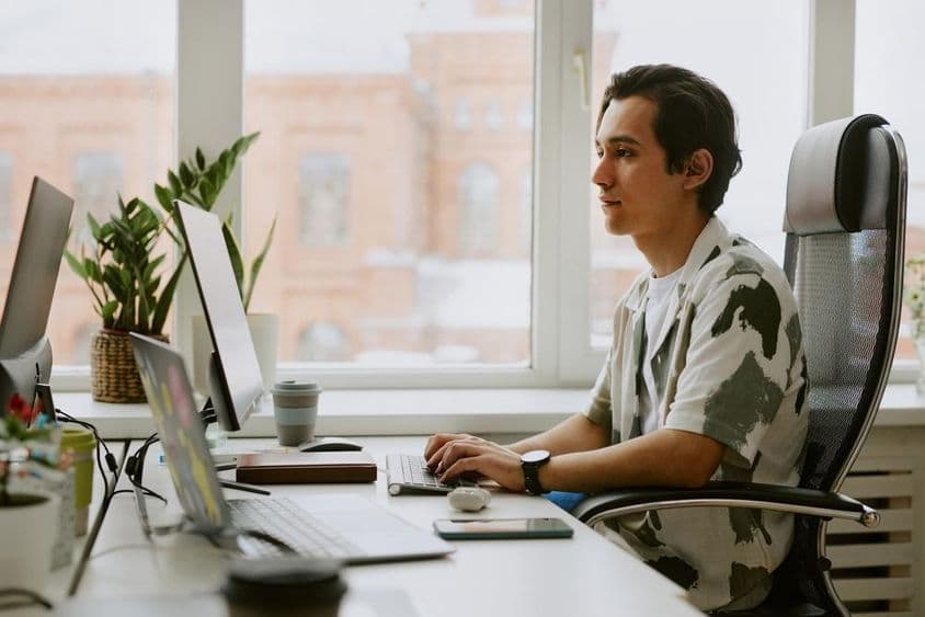 Jovem programador sentado à mesa, codificando em um computador em um escritório iluminado.