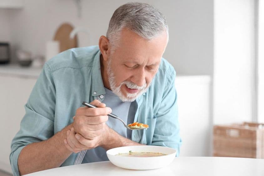 Homem idoso com Parkinson comendo em uma mesa de cozinha.
