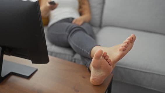 Uma jovem relaxando descalça em um sofá aconchegante na sala de estar, segurando um controle remoto.