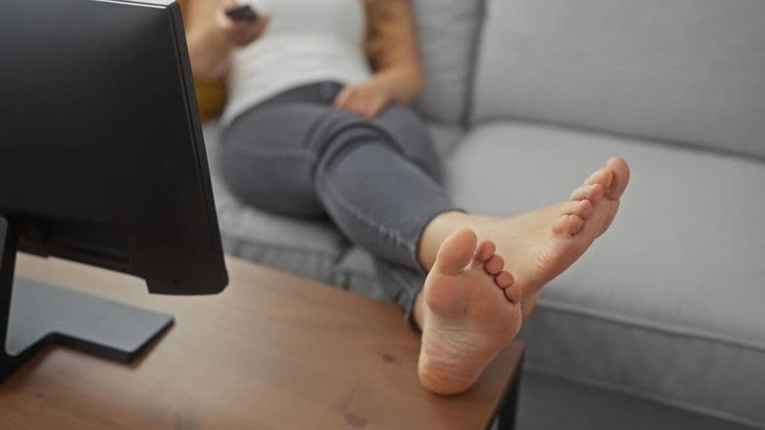 Uma jovem relaxando descalça em um sofá aconchegante na sala de estar, segurando um controle remoto.