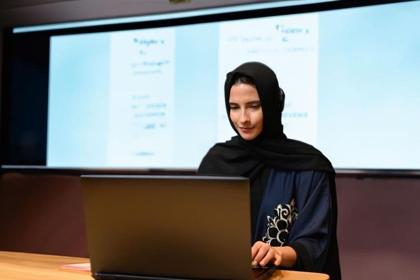 Professora usando Abaya Hijab com fundo de tela de projetor em uma sala de aula moderna com tela digital.