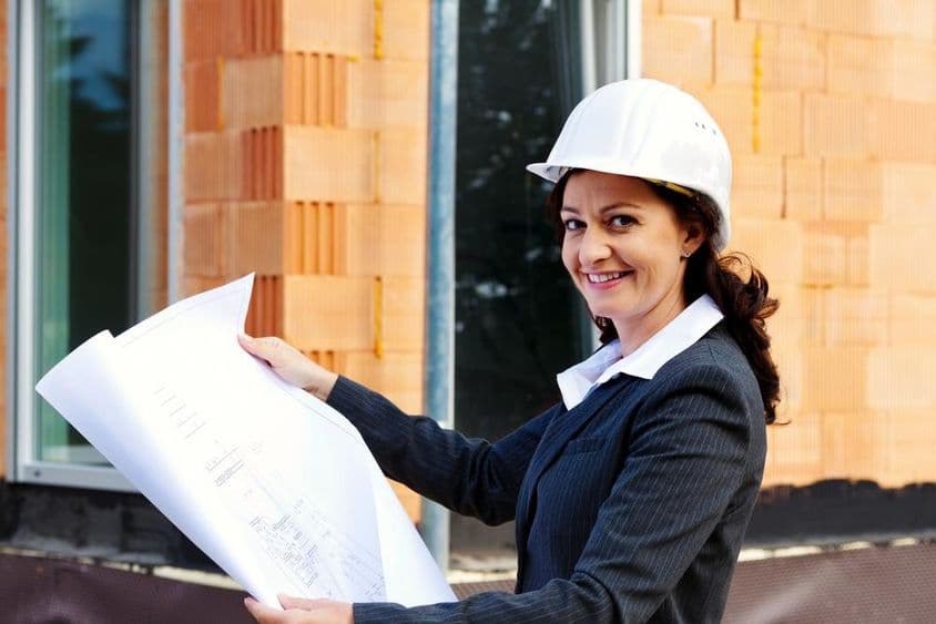 Arquiteta com planta em frente a uma casa em construção, usando capacete de segurança.