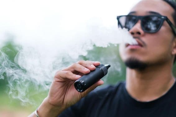 Homem asiático fumando com um cigarro eletrônico vaping, exalando fumaça pela boca e desfrutando ao ar livre no verão.