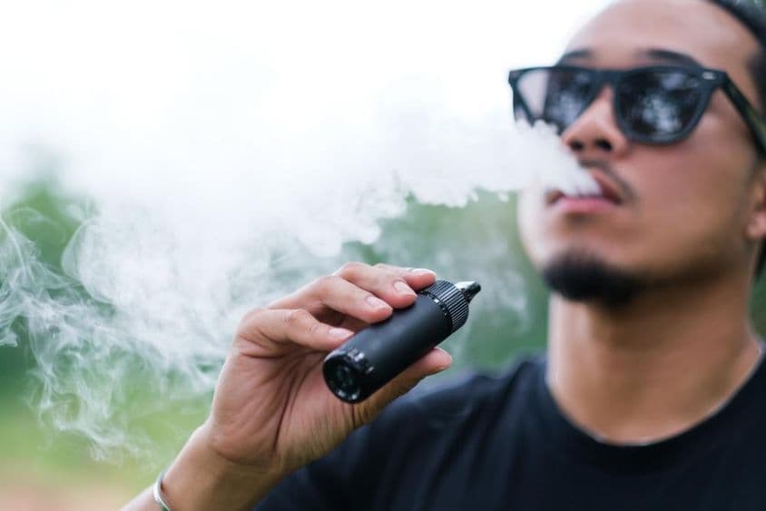 Homem asiático fumando com um cigarro eletrônico vaping, exalando fumaça pela boca e desfrutando ao ar livre no verão.