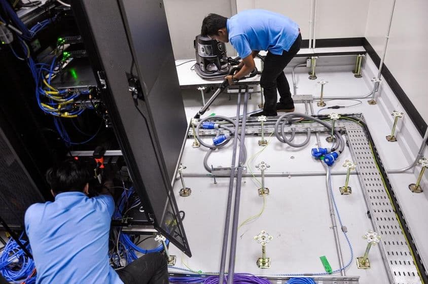 Trabalhadores construindo uma sala de servidores, piso elevado e gabinetes de rack.