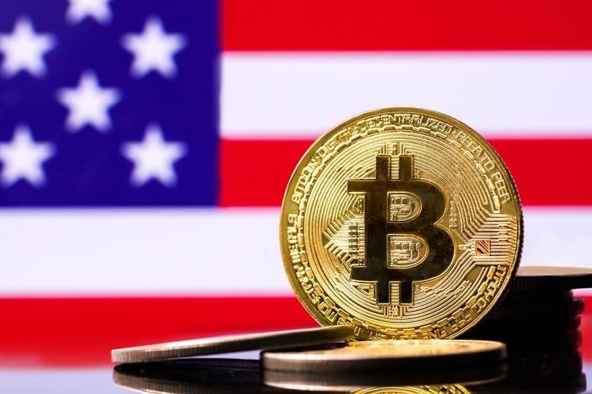 Moeda de metal Bitcoin dourada com a bandeira dos EUA ao fundo.