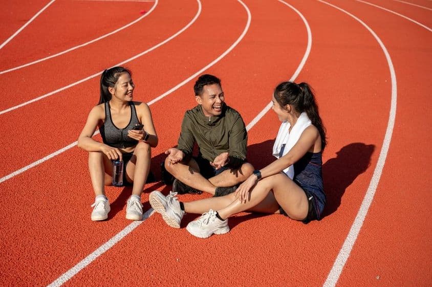 Um grupo de atletas sorridentes descansando em um campo esportivo sob o sol após o treino.