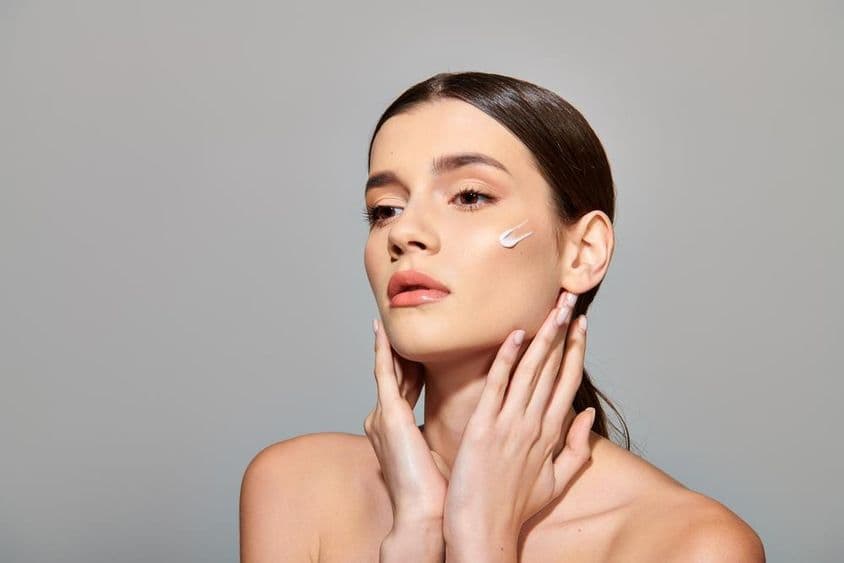 Uma jovem com cabelos castanhos aplica um creme de beleza no rosto, exalando uma sensação de autocuidado.