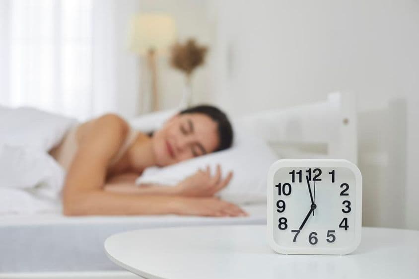 Despertador na mesa de cabeceira no quarto, com uma jovem mulher dormindo ao fundo.