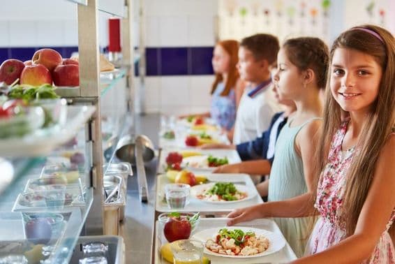 Menina sorridente com bandeja de comida, em fila com amigos durante o intervalo para o almoço na cantina escolar.