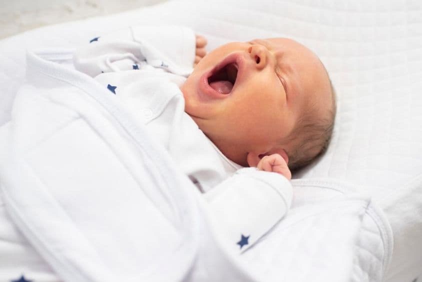 Bebê menino de uma semana bocejando em uma cama branca.
