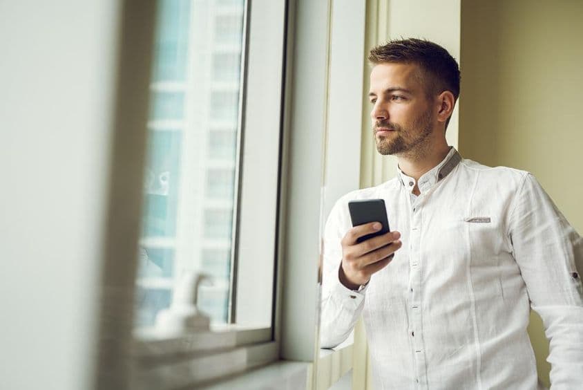 Empresário em pé perto de uma janela, segurando um smartphone.