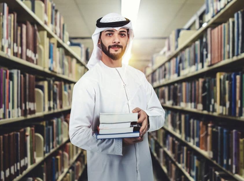 Estudante árabe em biblioteca segurando livros.
