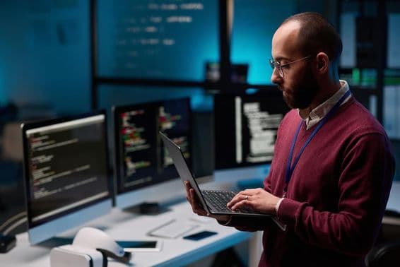 Programador de computador segurando um laptop aberto em um escritório de TI e cibersegurança.