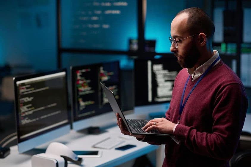 Programador de computador segurando um laptop aberto em um escritório de TI e cibersegurança.