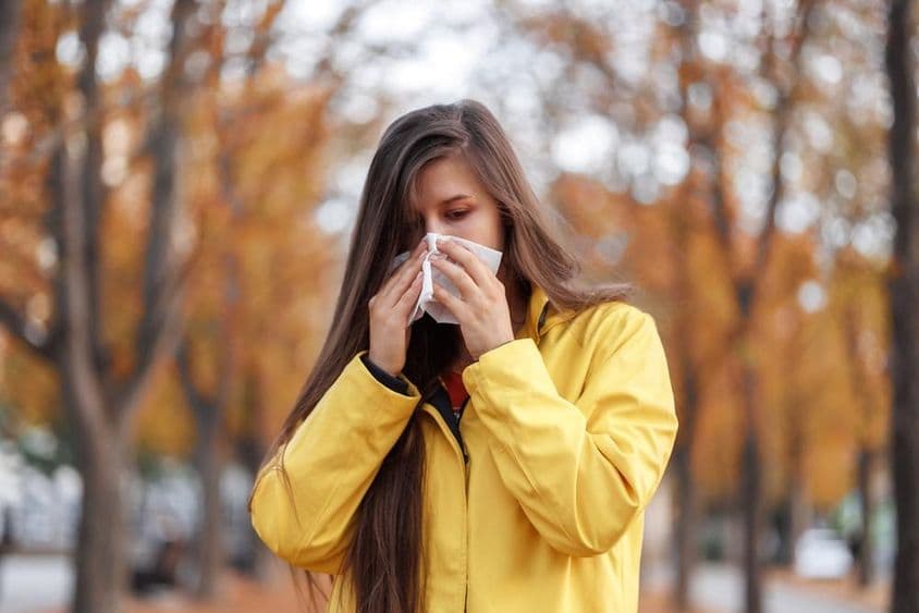 Mulher jovem em pé em um parque usando um casaco amarelo, segurando um lenço no nariz enquanto espirra.
