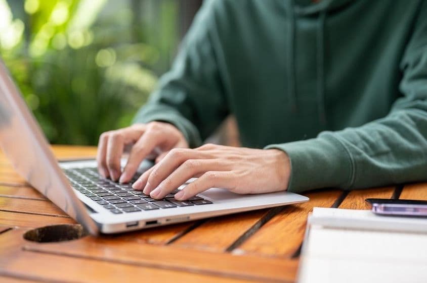Homem de suéter verde trabalhando remotamente em um laptop em uma mesa ao ar livre.