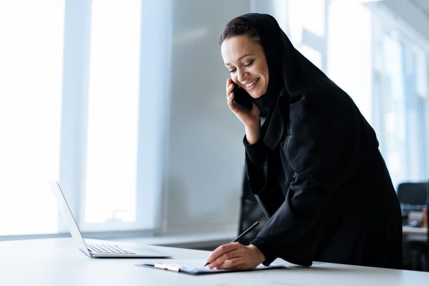 Uma bela mulher em uma abaya trabalhando no computador enquanto fala ao telefone.
