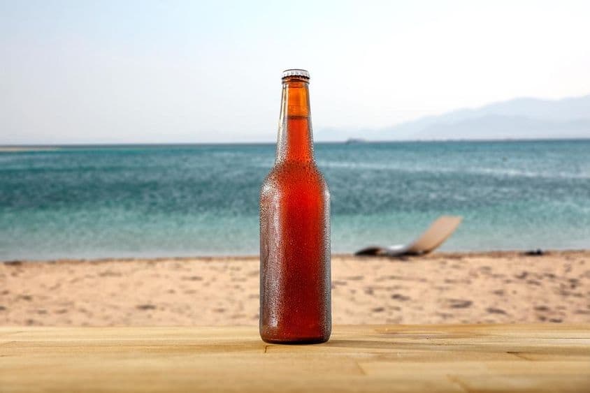 Uma garrafa de cerveja gelada sobre uma mesa de madeira na beira de uma praia arenosa.