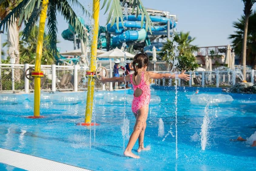 Menina pequena brincando em uma piscina com fontes em um parque aquático.