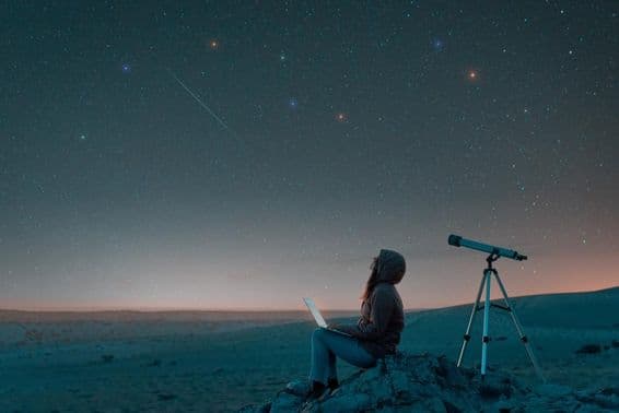 Mulher sentada no deserto com um laptop ao lado de um telescópio à noite, observando o céu estrelado.