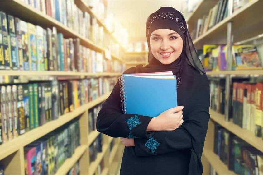 Estudante árabe em biblioteca com livros no braço.