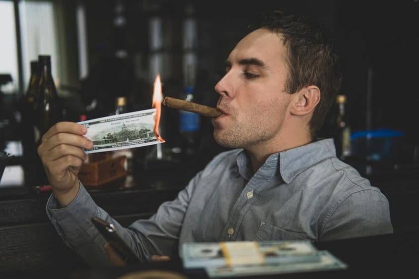 Jovem homem bonito acendendo charuto com cédula de cem dólares em um bar.