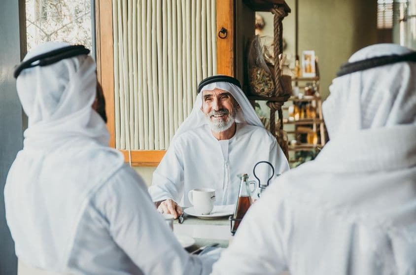 Três empresários apreciando chá em Dubai enquanto vestem trajes tradicionais dos Emirados.