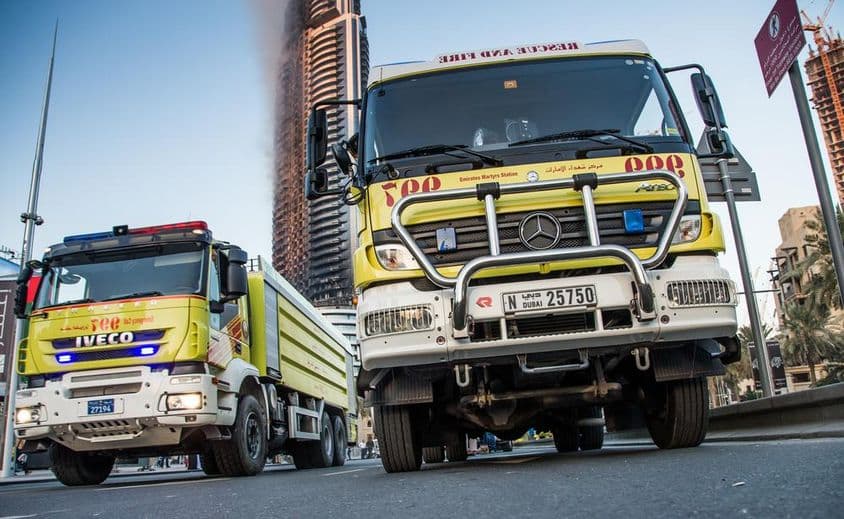 Caminhões de bombeiros de Dubai estacionados na rua, prédio em chamas ao fundo.