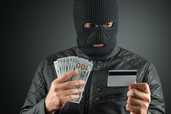 Criminoso com máscara de esqui segurando cartão de crédito contra um fundo escuro.