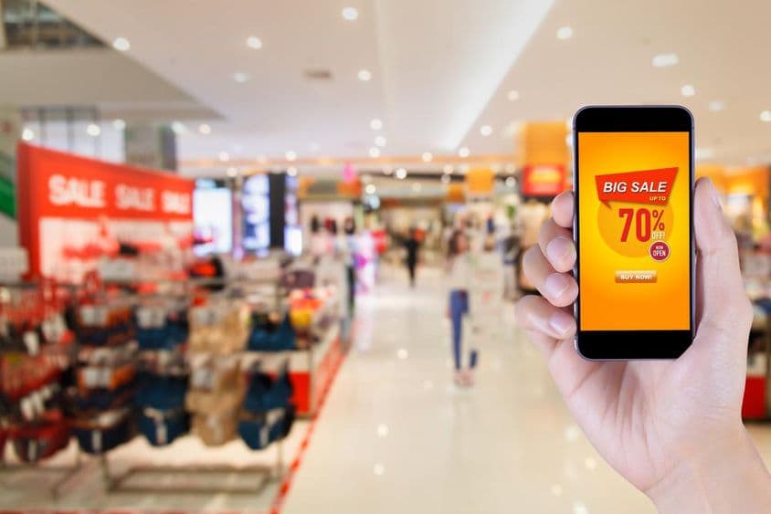 Smartphone exibindo tela de aplicativo de grande promoção com fundo borrado de shopping.