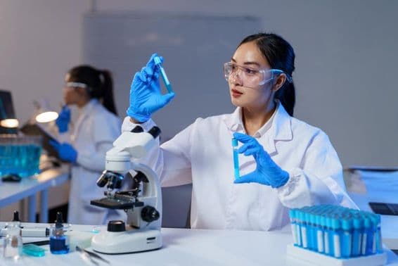 Cientista asiática de jaleco e luvas comparando dois tubos de ensaio.