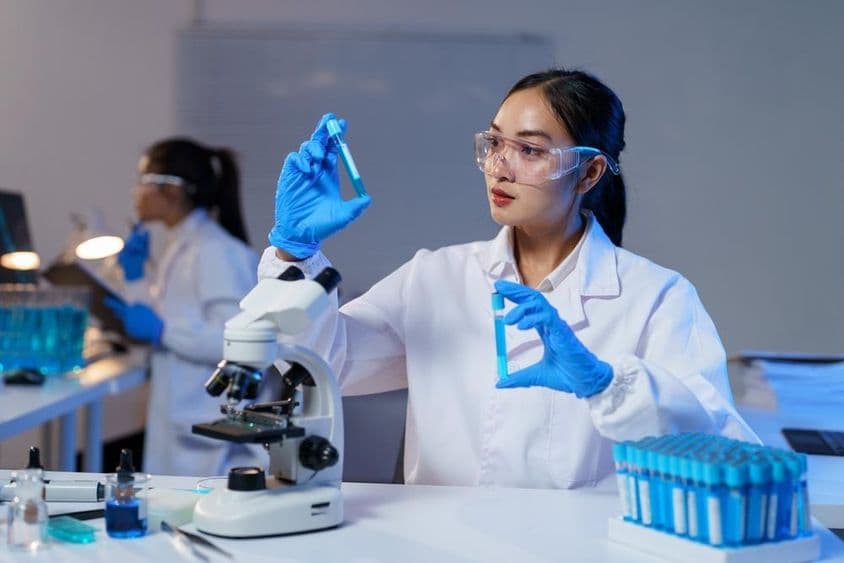 Cientista asiática de jaleco e luvas comparando dois tubos de ensaio.