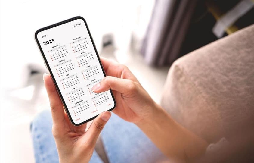 Mulher de negócios planejando atividades de 2025 com smartphone e calendário mensal.