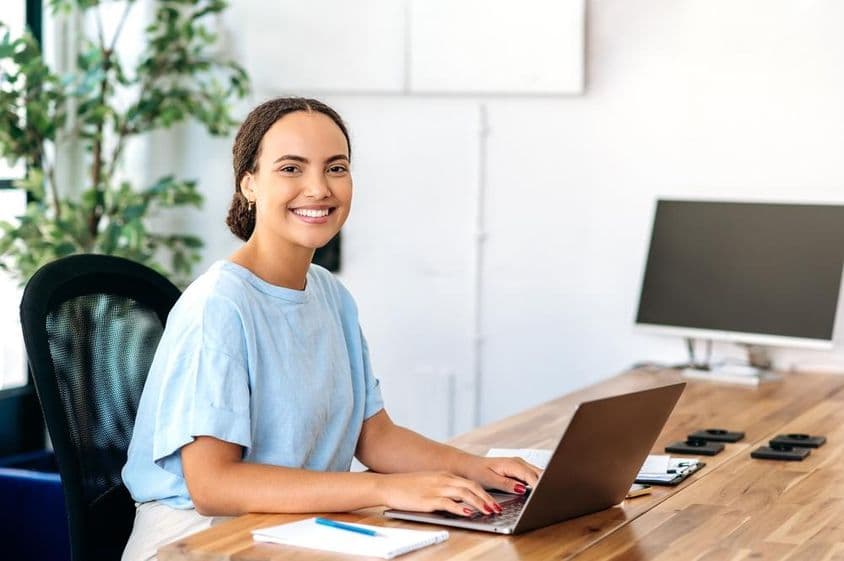 Mulher confiante sentada em uma mesa de escritório moderna, olhando para a câmera e sorrindo amigavelmente.
