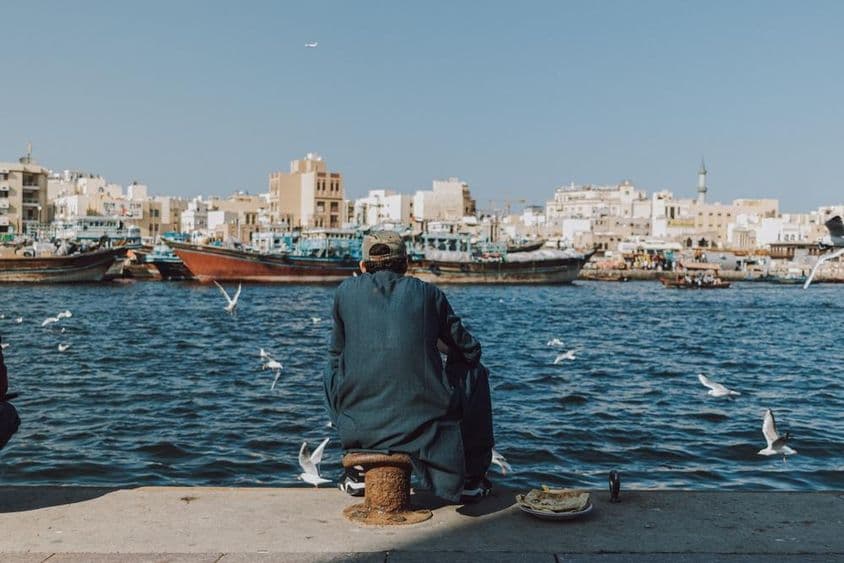 Homem desfrutando da vista à beira-mar com barcos e gaivotas no Dubai Creek.