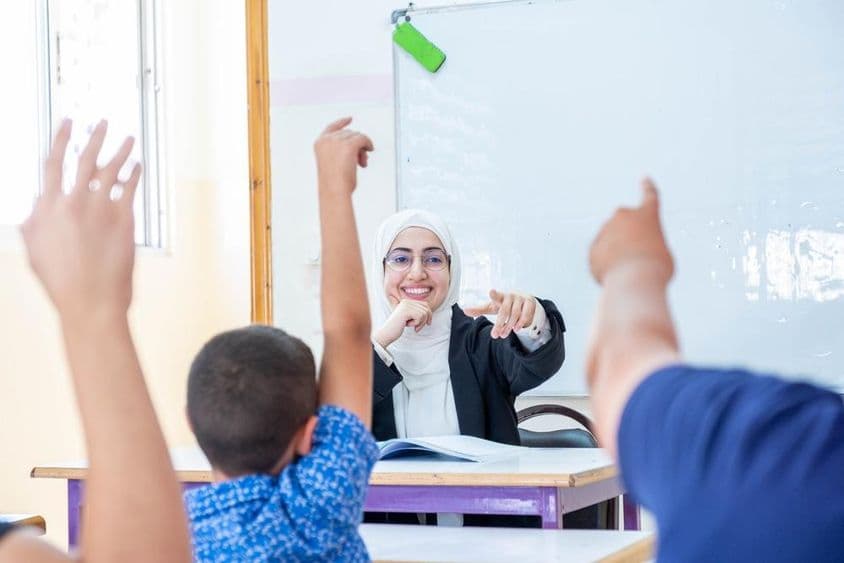 Estudantes levantando as mãos, indicando presença e respondendo ao professor.