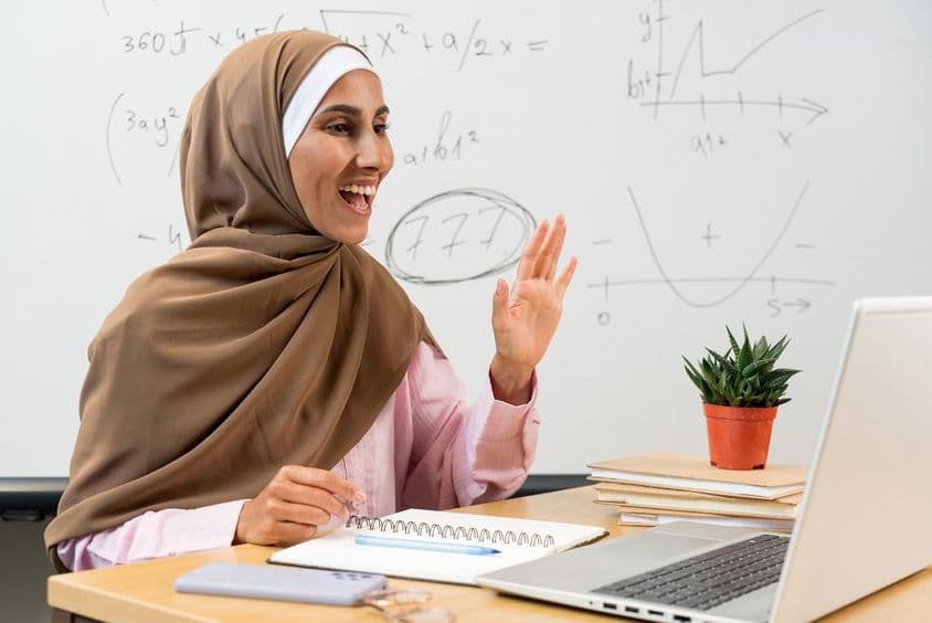 Professora em hijab acenando para estudantes durante uma aula online.